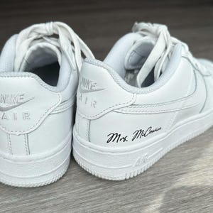 Könnte beinhalten: Ein Paar weiße Nike Air Sneaker mit weißen Schnürsenkeln. An der Ferse des rechten Schuhs befindet sich der schwarze, kursive Schriftzug Mrs. McCowin. Beide Schuhe zeigen das in Weiß gestickte Nike Air Logo auf der Fersenkappe und Air.