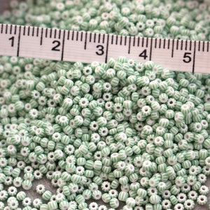 Könnte beinhalten: Ein Haufen kleiner, weißer Perlen mit grünen Streifen. Ein Lineal liegt über den Perlen und zeigt die Maße in Zoll an. Die Perlen sind zylindrisch mit einem Loch in der Mitte, zum Auffädeln geeignet. (ca. 2.5 cm)