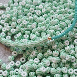 Könnte beinhalten: Nahaufnahme von weißen Perlen mit mintgrünen Streifen. Eine türkisfarbene Perlenkette mit goldenem Akzent ist ebenfalls zu sehen. Die Perlen sind verstreut, was auf ein Bastel- oder Schmuckthema hindeutet.