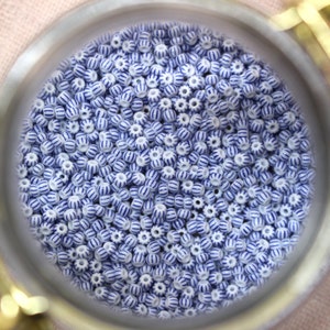 Puede incluir: Primer plano de una colecci&oacute;n de peque&ntilde;as cuentas blancas con rayas azules. Las cuentas son redondas con una textura ligeramente acanalada y est&aacute;n contenidas dentro de un recipiente circular met&aacute;lico. Las cuentas est&aacute;n dispersas, llenando el marco.