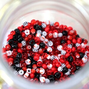 Könnte beinhalten: Ein Glasgefäß gefüllt mit roten, weißen und schwarzen Perlen. Die Perlen sind klein und rund und in einem zufälligen Muster angeordnet.