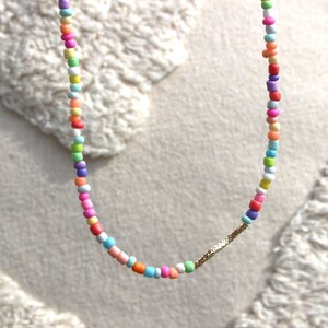 May include: A colorful beaded necklace with a gold bar accent. The beads are various shades of pink, blue, green, yellow, and white.