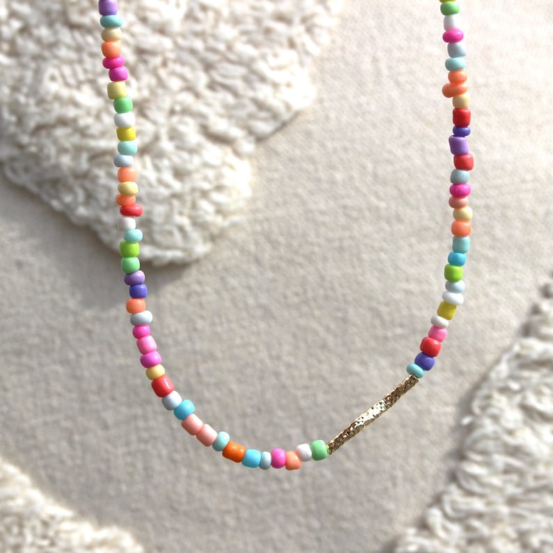 May include: A colorful beaded necklace with a gold bar accent. The beads are various shades of pink, blue, green, yellow, and white.
