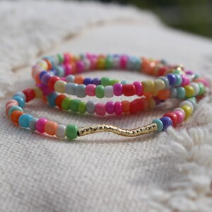 May include: Multicolored beaded bracelet with a gold wavy charm. The bracelet is made of small, colorful beads in a variety of shades, including pink, blue, green, yellow, and orange. The gold charm is a wavy line with a textured surface.
