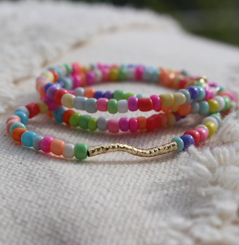May include: Multicolored beaded bracelet with a gold wavy charm. The bracelet is made of small, colorful beads in a variety of shades, including pink, blue, green, yellow, and orange. The gold charm is a wavy line with a textured surface.