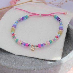 Könnte beinhalten: Ein farbenfrohes Perlenarmband mit einem rosa Kordelverschluss und einem goldenen Tropfenanhänger. Die Perlen sind in verschiedenen Schattierungen von Rosa, Blau, Gelb, Grün und Lila.
