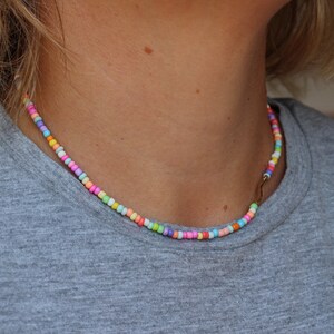 May include: A colorful beaded necklace with a gold clasp. The beads are small and round, and they are arranged in a random pattern. The necklace is worn by a person with a light grey shirt.