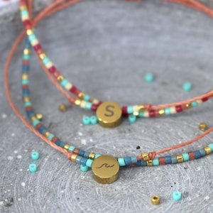 Könnte beinhalten: Zwei Armbänder mit Perlen und goldfarbenen runden Anhängern. Ein Anhänger hat den Buchstaben "S", der andere ein Wellensymbol. Die Armbänder haben eine Mischung aus farbigen Perlen in Rot-, Blau-, Türkis- und Goldtönen, auf einer korallenfarbenen Kordel. Schmuck.