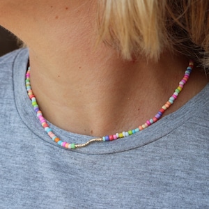 May include: A colorful beaded necklace with a gold chain accent. The beads are small and round, and they are arranged in a random pattern. The necklace is worn by a person with short blonde hair.