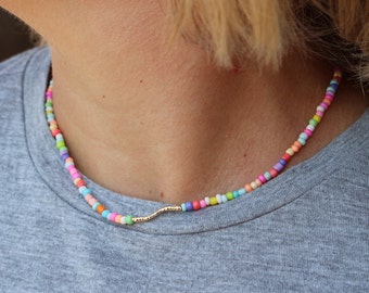 May include: A colorful beaded necklace with a gold chain accent. The beads are small and round, and they are arranged in a random pattern. The necklace is worn by a person with short blonde hair.