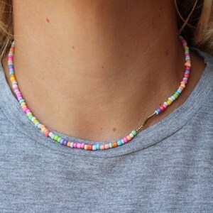 May include: A colorful beaded necklace with a gold chain accent. The beads are small and round, and they are arranged in a random pattern. The necklace is worn by a person, but the person's face is not visible.