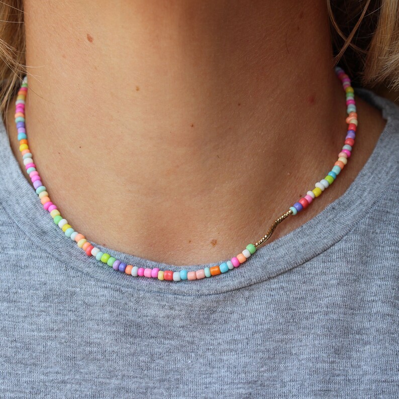 May include: A colorful beaded necklace with a gold chain accent. The beads are small and round, and they are arranged in a random pattern. The necklace is worn by a person, but the person's face is not visible.