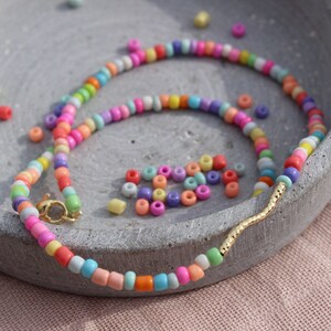 May include: A colorful beaded necklace with a gold accent. The necklace is made of small, round beads in various shades of pink, blue, green, yellow, and orange. The beads are strung on a thin, clear thread. The necklace has a gold clasp and a gold accent in the shape of a wavy line.