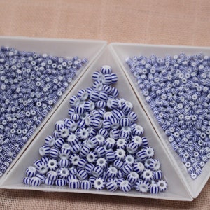 Puede incluir: Cuentas a rayas blancas y azules dispuestas en tres bandejas triangulares. Las cuentas tienen forma cil&iacute;ndrica con una serie de rayas azules alrededor de la circunferencia. Estas cuentas son un suministro artesanal, adecuado para la fabricaci&oacute;n de joyas u otros proyectos creativos.