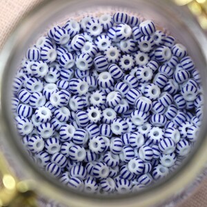 Puede incluir: Primer plano de un tarro de cristal lleno de numerosas cuentas blancas adornadas con rayas azules. Las cuentas son cil&iacute;ndricas con un agujero en el centro, creando un patr&oacute;n de colores alternos. Las cuentas son probablemente para la fabricaci&oacute;n de joyas.
