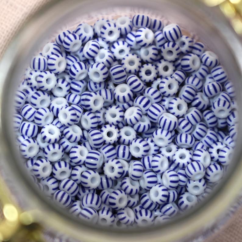 Puede incluir: Primer plano de un tarro de cristal lleno de numerosas cuentas blancas adornadas con rayas azules. Las cuentas son cil&iacute;ndricas con un agujero en el centro, creando un patr&oacute;n de colores alternos. Las cuentas son probablemente para la fabricaci&oacute;n de joyas.