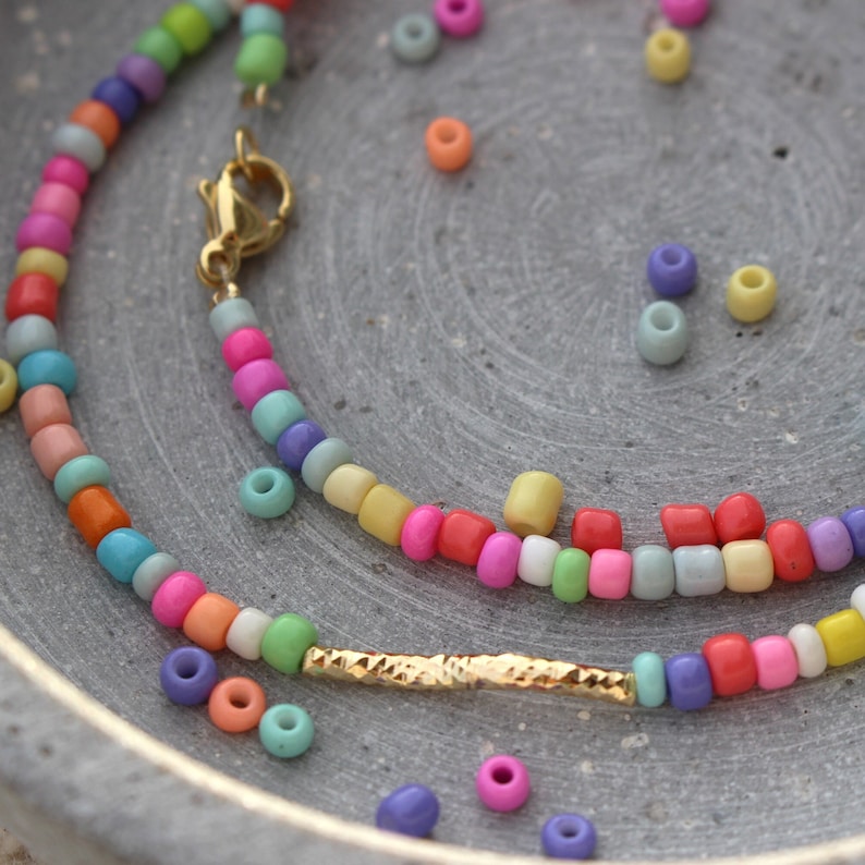 May include: A colorful beaded necklace with a gold clasp and a gold bar charm. The beads are in various shades of pink, blue, green, yellow, and orange. The necklace is laid out on a gray surface with loose beads scattered around it.