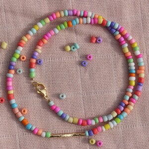 May include: A colorful beaded necklace with a gold clasp and a gold bar accent. The necklace is made of small, round beads in various shades of pink, blue, green, yellow, and orange. The beads are strung on a clear thread and the necklace is lying on a pink fabric background.