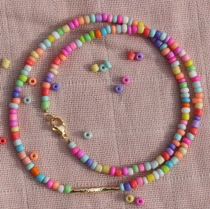 May include: A colorful beaded necklace with a gold clasp and a gold bar accent. The necklace is made of small, round beads in various shades of pink, blue, green, yellow, and orange. The beads are strung on a clear thread and the necklace is lying on a pink fabric background.