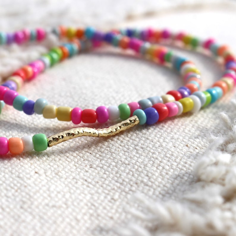 May include: A colorful beaded necklace with a gold bar accent. The beads are in various shades of pink, blue, green, yellow, and white.