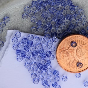 Könnte beinhalten: Nahaufnahme von zahlreichen kleinen, runden Perlen mit transparentem Grund und blau-weißem Streifenmuster. Eine kupferne Euro-Münze liegt neben den Perlen als Größenvergleich. Die Perlen sind auf einem weißen Tuch und einer grauen Oberfläche verstreut.