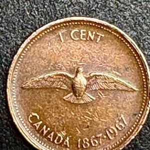 Puede incluir: Una moneda canadiense de un centavo de color cobre. La moneda presenta un pájaro con las alas extendidas, el texto "1 CENT" y las palabras "CANADA 1867-1967". La moneda tiene un diseño detallado y forma circular.