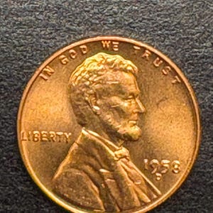 Puede incluir: Una moneda de cobre Lincoln de 1958. El anverso presenta un perfil de Abraham Lincoln, la palabra "LIBERTY" y la frase "IN GOD WE TRUST". La moneda tiene el número "00120" en la esquina superior izquierda.