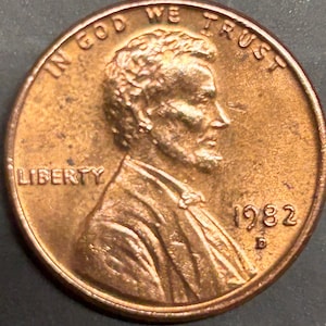 Puede incluir: Un centavo estadounidense de cobre de 1982 con la inscripción "In God We Trust" y "Liberty" en el anverso. La moneda presenta un perfil de Abraham Lincoln.