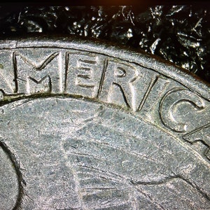 May include: Close-up of a silver coin with the word "AMERICA" arched across the top. The coin's surface shows intricate details and textures, with a blurred background.