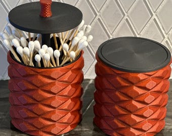 May include: Two red, geometric-patterned containers with black lids. One holds cotton swabs, the other is empty. The containers are cylindrical and have a textured, diamond-like pattern. The lid of the container with swabs has a small, red knob.
