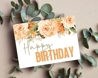 May include: A white birthday card with the words "Happy Birthday" in green and orange. The card features peach-coloured roses and green foliage. Eucalyptus branches surround the card on a neutral background.