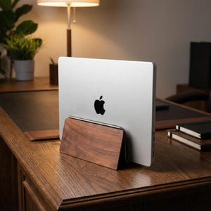 May include: A silver laptop with a black Apple logo is held by a wooden stand. The stand is made of dark brown wood and has a rectangular shape. The laptop and stand are on a wooden desk.
