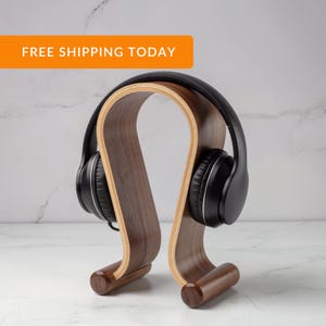 Peut inclure: Un support pour casque audio en bois avec un design incurvé. Le support est en bois de noyer et a une finition naturelle. Un casque audio noir est posé sur le support.