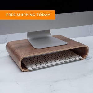 May include: A curved wooden monitor stand supporting a silver computer monitor. A white keyboard is placed on the shelf below. The stand is made of light brown wood and sits on a white marble surface.