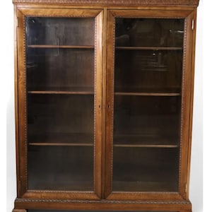 Op de afbeelding: Een lichtbruine houten boekenkast met twee glazen deuren. De boekenkast heeft meerdere planken en decoratieve versieringen langs de boven- en onderkant. De binnenkant van de kast is donker. De deuren zijn van hout.