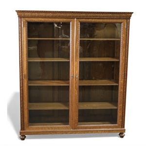 May include: A wooden bookcase with two glass-paned doors. The bookcase has multiple shelves and is made of light brown wood. The doors have a decorative trim around the edges. The bookcase is a classic piece of furniture.
