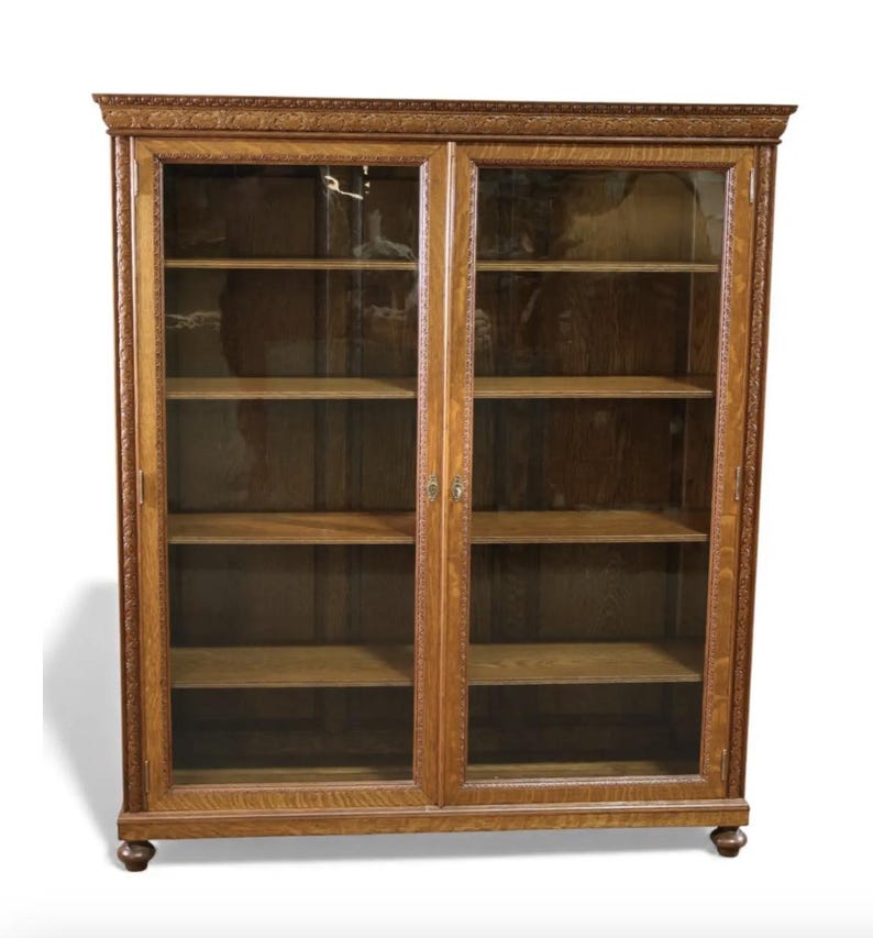 May include: A wooden bookcase with two glass-paned doors. The bookcase has multiple shelves and is made of light brown wood. The doors have a decorative trim around the edges. The bookcase is a classic piece of furniture.