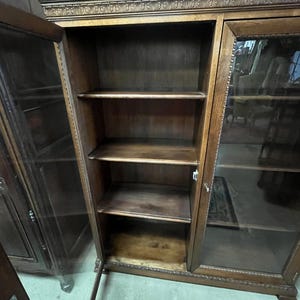Op de afbeelding: Een donkerhouten boekenkast met glazen deuren en drie planken. De boekenkast heeft sierlijke gravures langs de bovenkant en rond de glazen deurkozijnen. De planken zijn leeg en de deuren staan open.