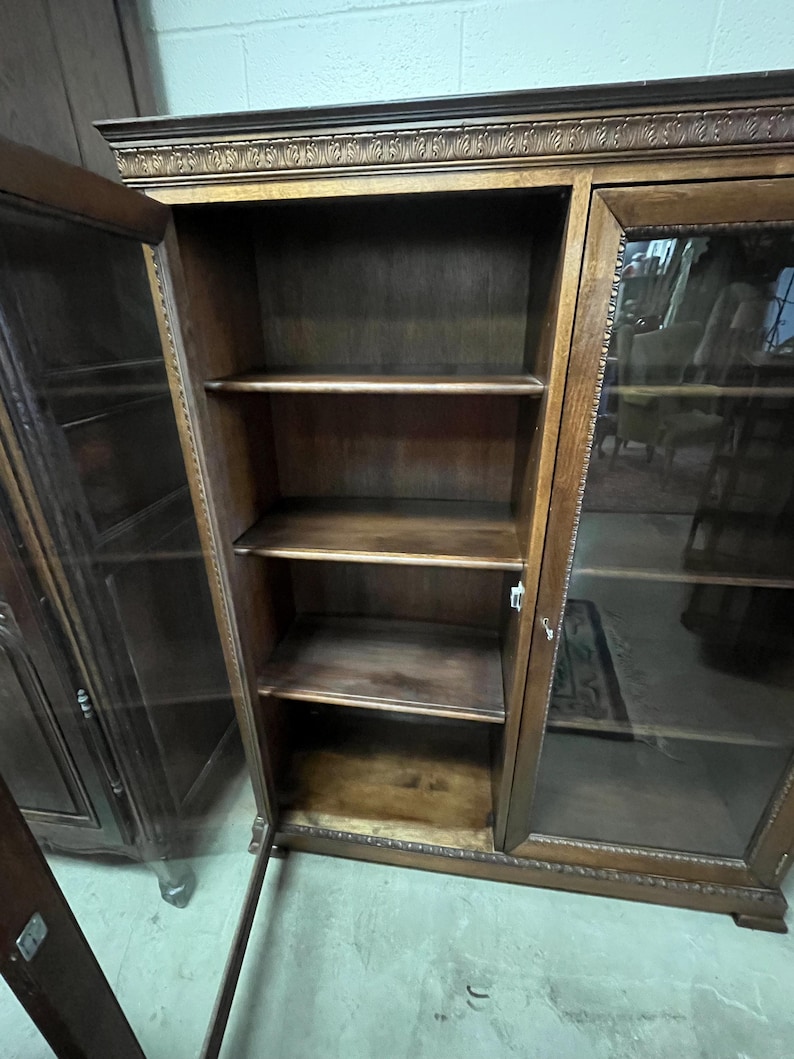 Op de afbeelding: Een donkerhouten boekenkast met glazen deuren en drie planken. De boekenkast heeft sierlijke gravures langs de bovenkant en rond de glazen deurkozijnen. De planken zijn leeg en de deuren staan open.