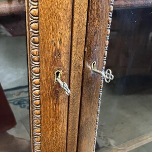 Op de afbeelding: Close-up van een houten kastdeur met twee sleutels in de sleutelgaten. Het hout heeft een rijke, warme bruine tint met zichtbare nerf. Het deurkozijn heeft een decoratieve, gebeeldhouwde rand. De kast heeft glazen panelen.
