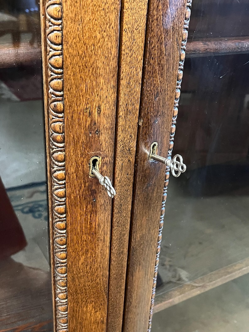 Op de afbeelding: Close-up van een houten kastdeur met twee sleutels in de sleutelgaten. Het hout heeft een rijke, warme bruine tint met zichtbare nerf. Het deurkozijn heeft een decoratieve, gebeeldhouwde rand. De kast heeft glazen panelen.