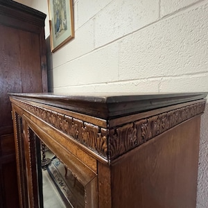 Op de afbeelding: Een donkerbruine houten kast met glazen deuren en sierlijke gesneden details langs de bovenrand. Een ingelijste foto hangt aan de witte bakstenen muur achter de kast.