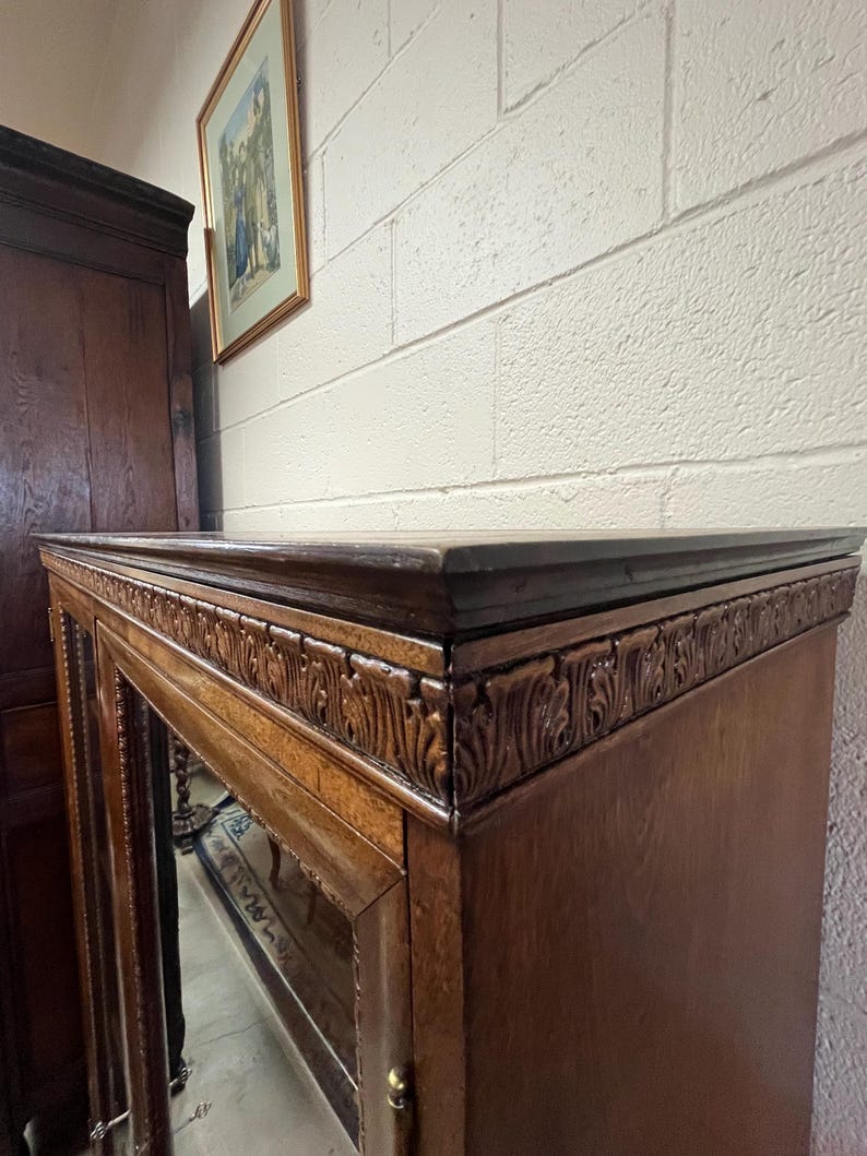 Op de afbeelding: Een donkerbruine houten kast met glazen deuren en sierlijke gesneden details langs de bovenrand. Een ingelijste foto hangt aan de witte bakstenen muur achter de kast.