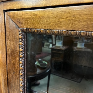 Op de afbeelding: Close-up van een houten kastdeur met een glazen paneel. Het deurkozijn is een bruine kleur met gesneden details langs de randen. Het glazen paneel weerspiegelt de binnenkant van een kamer, inclusief meubels.