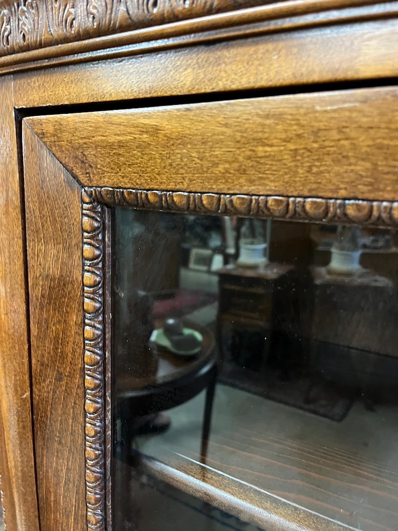 Op de afbeelding: Close-up van een houten kastdeur met een glazen paneel. Het deurkozijn is een bruine kleur met gesneden details langs de randen. Het glazen paneel weerspiegelt de binnenkant van een kamer, inclusief meubels.