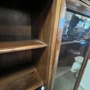 Op de afbeelding: Een donkerbruine houten kast met glazen deur en planken. De kast heeft een gesneden ontwerp langs de bovenkant en het deurkozijn. De planken zijn zichtbaar in de kast. De glazen deur weerspiegelt de omgeving.