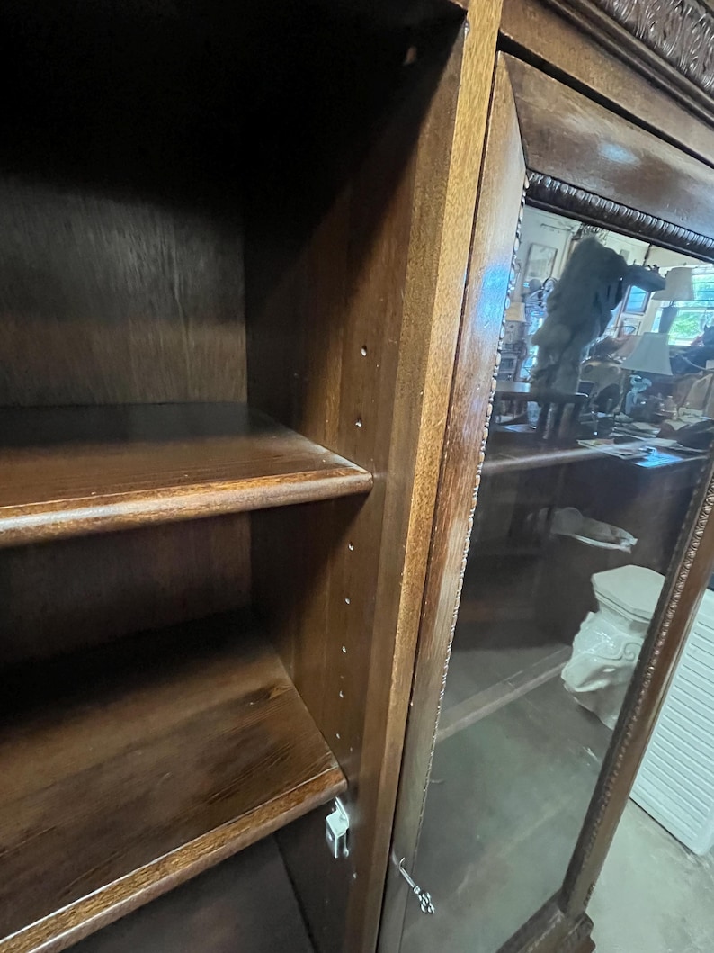 Op de afbeelding: Een donkerbruine houten kast met glazen deur en planken. De kast heeft een gesneden ontwerp langs de bovenkant en het deurkozijn. De planken zijn zichtbaar in de kast. De glazen deur weerspiegelt de omgeving.