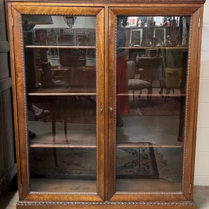 Op de afbeelding: Een hoge, antieke houten kast met twee glazen deuren. De kast heeft een warme bruine kleur met decoratieve sierlijsten langs de bovenkant, onderkant en rond het glas. De binnenkant heeft planken en de glazen deuren weerspiegelen de kamer.