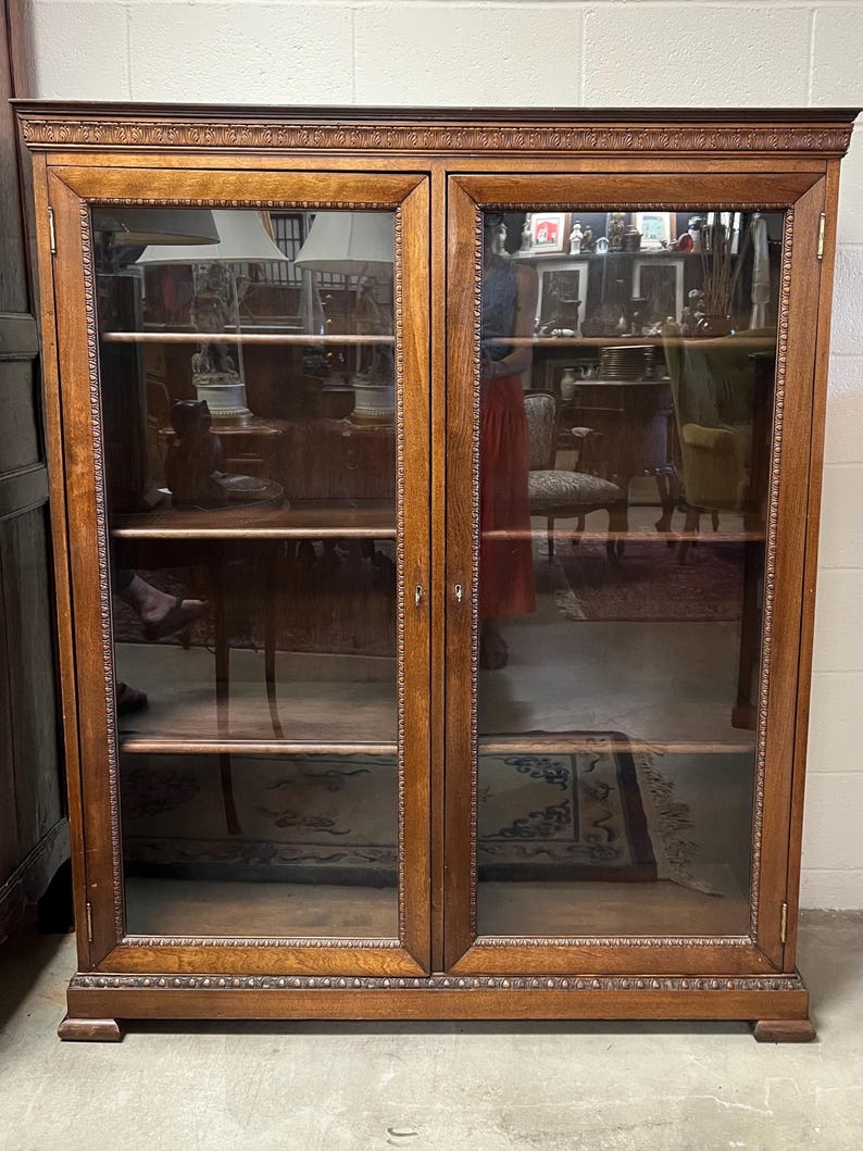 Op de afbeelding: Een hoge, antieke houten kast met twee glazen deuren. De kast heeft een warme bruine kleur met decoratieve sierlijsten langs de bovenkant, onderkant en rond het glas. De binnenkant heeft planken en de glazen deuren weerspiegelen de kamer.