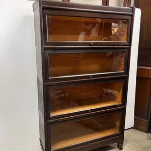 May include: A dark wood barrister bookcase with four glass-fronted sections. The bookcase has a rectangular shape and sits on short legs. The glass doors have small, round knobs. The interior of the bookcase is visible through the glass.